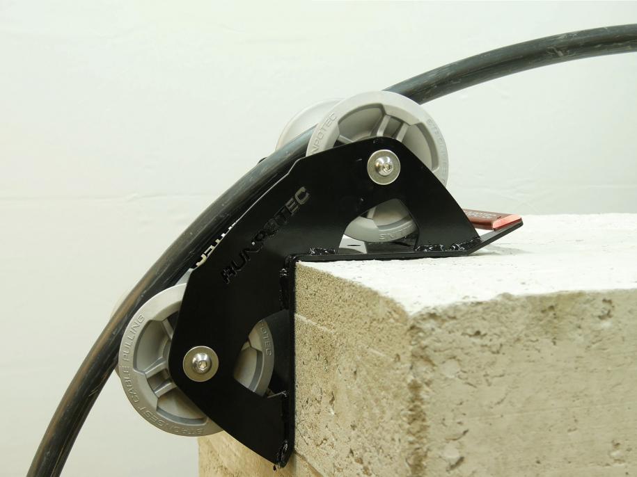 A 90 degree cable guide roller with a robust metal frame and heavy-duty aluminium rollers designed to pull cables gently around corners. Example photo showing roller attached to the corner of a concrete block.